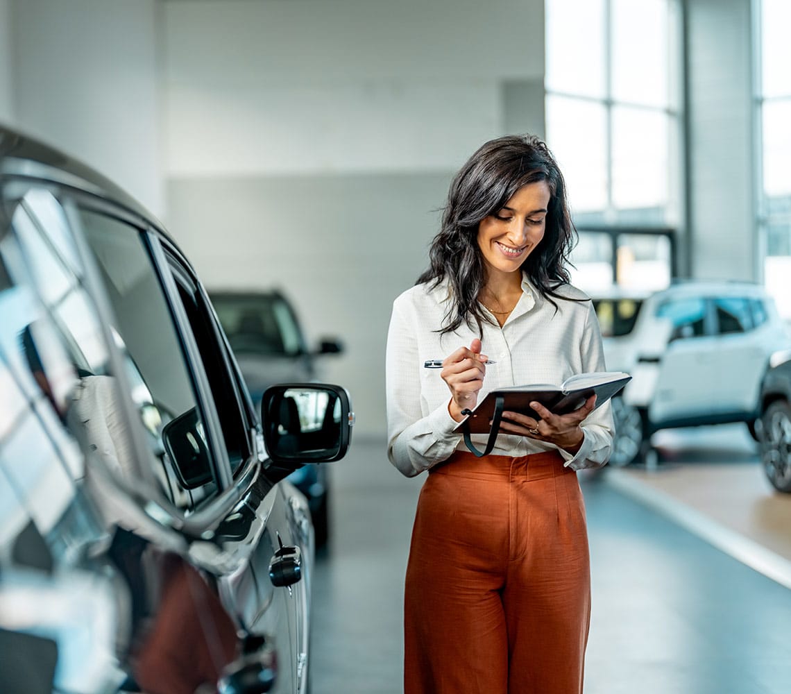 A smiling woman at a car dealership diligently writing notes on a clipboard, surrounded by various vehicles on display.